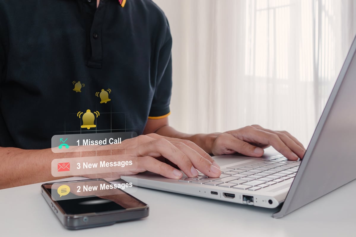 Man working on a laptop while notifications of missed calls and messages arrive on his smartphone